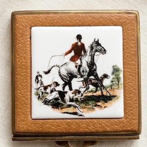 May include: A small, square trinket box with a tan leather exterior and a brass frame. The center features a white tile with a hunting scene illustration in shades of brown, black, and green. The illustration depicts a rider on horseback with hounds.