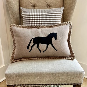 May include: A brown linen armchair with a brown and white checkered pillow and a beige linen pillow with a black horse silhouette. The chair is sitting on a woven rug.