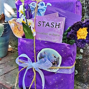 May include: A purple gift box wrapped in matching tissue paper, tied with a white ribbon and twine. The box contains a tea bag labeled "STASH Breakfast in Paris", flowers, and a crocheted purple and yellow flower.