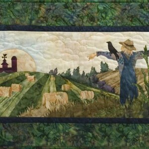 May include: A quilted wall hanging depicting a scarecrow standing in a field of corn with a crow perched on its outstretched arm. The scarecrow is wearing a straw hat and overalls. In the background, a farmhouse and a field of hay bales are visible. The sky is cloudy and the sun is setting.