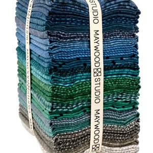 May include: A stack of fabric squares in shades of blue, teal, and gray, tied with a cream ribbon printed with "MAYWOOD STUDIO". The fabric has a textured appearance, with variations in color and pattern. The stack is neatly arranged, showcasing the different hues.