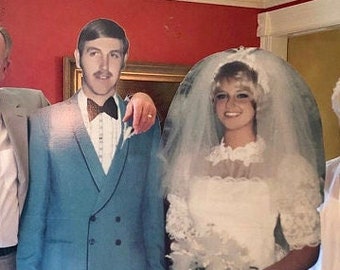 May include: A life-sized cardboard cutout of a wedding couple. The person on the left wears a teal double-breasted tuxedo jacket with a black bowtie. The person on the right wears a white lace wedding dress, a veil, and holds a white bouquet.