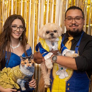 Puede incluir: Una pareja vestida como Bella y la Bestia de La Bella y la Bestia, con sus mascotas vestidas como los mismos personajes. La mujer sostiene un gato vestido como Bella, y el hombre sostiene un perro vestido como la Bestia.