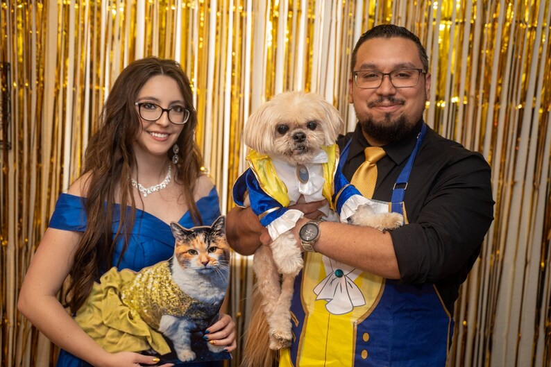 Puede incluir: Una pareja vestida como Bella y la Bestia de La Bella y la Bestia, con sus mascotas vestidas como los mismos personajes. La mujer sostiene un gato vestido como Bella, y el hombre sostiene un perro vestido como la Bestia.