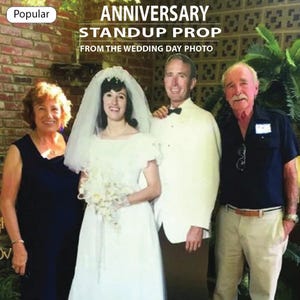 May include: A standup prop featuring a photo of a wedding party. The text at the top reads "ANNIVERSARY STANDUP PROP FROM THE WEDDING DAY PHOTO". The photo shows a bride, groom, and two other people. The background is a brick wall and greenery.