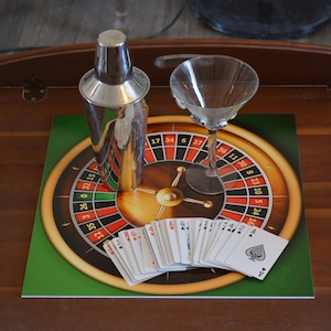 May include: A silver metal cocktail shaker and a clear glass martini glass placed on a square mat featuring a printed roulette wheel graphic. A fanned-out deck of standard playing cards is positioned in front of the glass and shaker on the mat.
