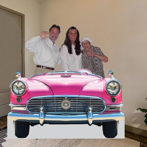 May include: Three people stand in front of a pink vintage car. The car has chrome bumpers and headlights. The people are smiling and appear to be having fun.