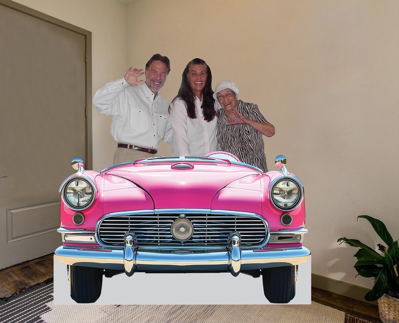 May include: Three people stand in front of a pink vintage car. The car has chrome bumpers and headlights. The people are smiling and appear to be having fun.