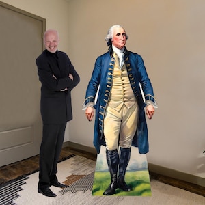 May include: A cardboard cutout of George Washington, the first president of the United States, wearing a blue coat with gold trim and white pants. He is standing on a green grassy field with a blue sky in the background.