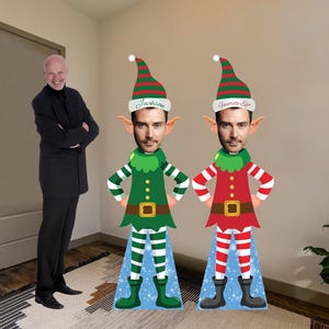May include: Two cardboard cutouts of elves, one green and one red, stand side-by-side. Each elf has a face, a green and red striped hat, and a matching outfit. The green elf has the name "Jackson" on its hat. A man in a black suit stands to the left.