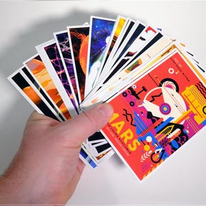 May include: A hand holds a stack of colorful postcards with various designs. The top card features a red background with a colorful illustration of a futuristic city and the text "Mars: A New Frontier for Adventure".