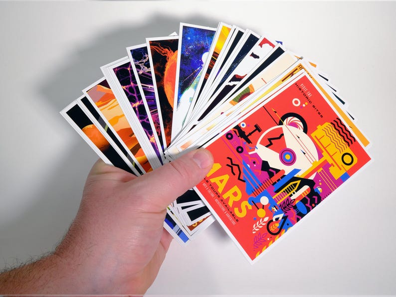 May include: A hand holds a stack of colorful postcards with various designs. The top card features a red background with a colorful illustration of a futuristic city and the text "Mars: A New Frontier for Adventure".