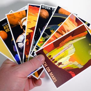 May include: A hand holds a stack of colorful postcards with images of Mars and astronauts. The text on the postcards includes "Work the Crater", "Some Assembly Required", "Technicians", and "Reach on Mars".