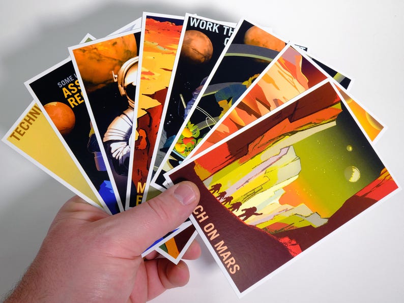May include: A hand holds a stack of colorful postcards with images of Mars and astronauts. The text on the postcards includes "Work the Crater", "Some Assembly Required", "Technicians", and "Reach on Mars".