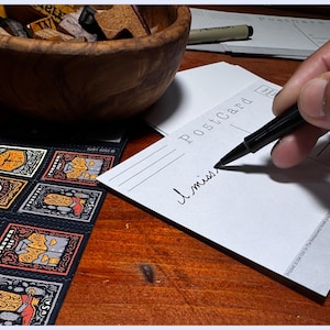 May include: A white postcard with the word "Postcard" printed on it. Someone is writing "I miss" on the postcard with a black pen.