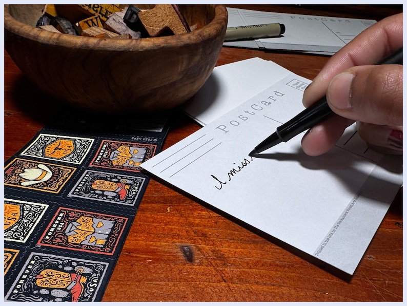 May include: A white postcard with the word "Postcard" printed on it. Someone is writing "I miss" on the postcard with a black pen.