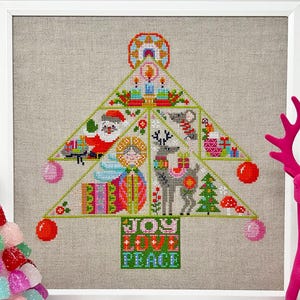 Puede incluir: Diseño de árbol de Navidad bordado a punto de cruz con Papá Noel, renos y regalos. El árbol es verde con adornos rojos y rosas. Las palabras "JOY LOVE PEACE" están bordadas en la base. La obra está enmarcada en un marco blanco.