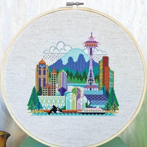 May include: A colorful cross-stitch embroidery hoop depicting a stylized cityscape with a mountain, a skyscraper, and a space needle. The embroidery is on a white fabric background.