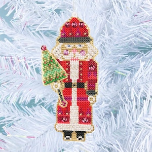 Puede incluir: Un adorno navideño rojo y dorado que representa a un cascanueces sosteniendo un pequeño árbol de Navidad verde. El cascanueces lleva un uniforme rojo y dorado con un sombrero negro y ribetes blancos. El adorno está decorado con lentejuelas y cuentas.