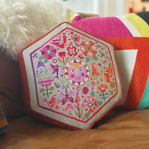 Peut inclure: Un coussin hexagonal avec un motif brodé coloré représentant un soleil, des oiseaux, des fleurs et des papillons. Le coussin est en tissu beige clair et a une bordure rouge.