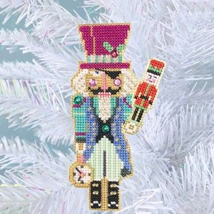 May include: A colorful, hand-stitched Christmas ornament featuring a nutcracker with a pink hat, a blue and green jacket, and a gold pocket watch. The nutcracker is holding a smaller, red nutcracker.