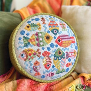 May include: A circular cushion with a cross-stitch design of colourful fish. The fish are in shades of yellow, orange, green, and pink, set against a white background with blue accents. The cushion has a green border.
