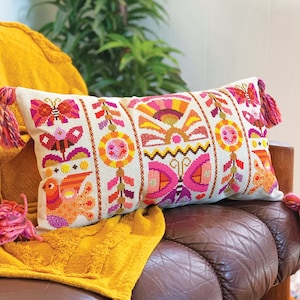 May include: A colourful embroidered cushion with a geometric pattern featuring butterflies, flowers, and suns. The cushion has tassels on each end and is resting on a brown leather sofa.