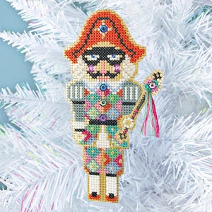 May include: A colorful, beaded nutcracker ornament with a red hat, black eye mask, and a gold and silver patterned uniform. The ornament has a gold tassel and a beaded stick.