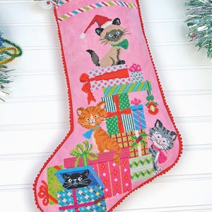May include: Pink Christmas stocking with a red scalloped trim, featuring embroidered cats wearing Santa hats and bows, surrounded by colourful gift boxes. The stocking is decorated with festive holiday accents.
