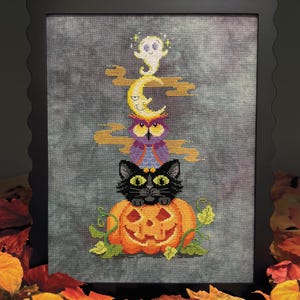May include: A cross-stitch embroidery in a black scalloped frame featuring a vertical stack of a jack-o'-lantern, a black cat, a purple owl, a yellow crescent moon, and a small white ghost, all set against a dark gray mottled fabric background.