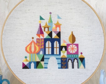 May include: A colorful cross-stitch embroidery of a castle with geometric shapes, framed in a wooden hoop and displayed on an easel. The embroidery is surrounded by yarn balls and other craft supplies.