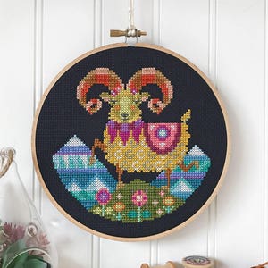 Könnte beinhalten: Ein Kreuzstich-Stickrahmen mit schwarzem Hintergrund, der ein farbenfrohes Widder mit einem rosa, lila und gelben Muster zeigt. Der Widder steht auf einer grünen, blauen und weißen Berglandschaft mit Blumen.