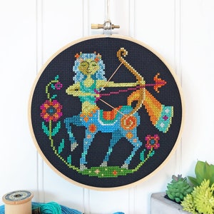 May include: A colorful cross-stitch embroidery hoop with a depiction of the Sagittarius zodiac sign. The Sagittarius is a centaur with a bow and arrow, surrounded by flowers. The embroidery is on black fabric.