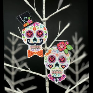Puede incluir: Dos adornos de calavera de azúcar coloridos con ojos negros, corazones rosas y detalles florales. La calavera de la izquierda lleva un sombrero de copa negro con una corbata de lazo. La calavera de la derecha lleva un velo blanco y una flor rosa.