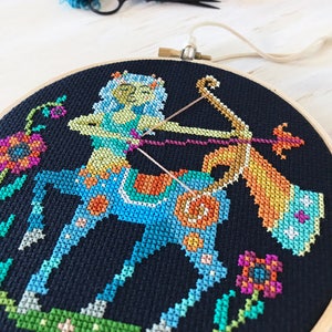 May include: A colorful cross-stitch embroidery hoop featuring a Sagittarius zodiac sign with a blue and green centaur holding a bow and arrow. The background is black with colorful flowers.