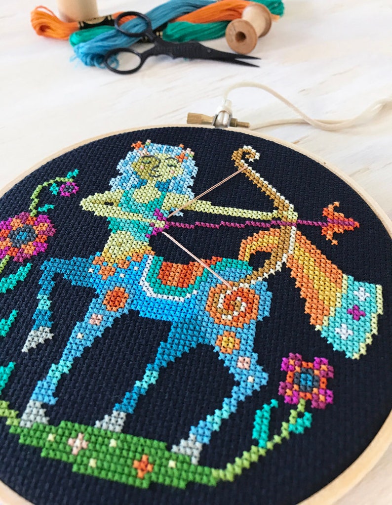 May include: A colorful cross-stitch embroidery hoop featuring a Sagittarius zodiac sign with a blue and green centaur holding a bow and arrow. The background is black with colorful flowers.