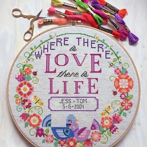 K&ouml;nnte beinhalten: Ein runder Stickrahmen mit wei&szlig;em Hintergrund und bunter Blumenstickerei. Die Stickerei zeigt den Text "Where there is love there is life" mit einer personalisierten Inschrift darunter: "Jess + Tom 5.15.2004".