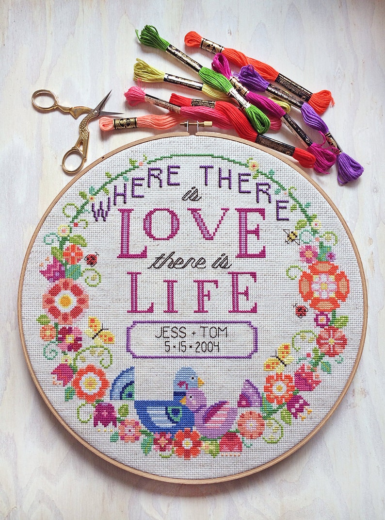 K&ouml;nnte beinhalten: Ein runder Stickrahmen mit wei&szlig;em Hintergrund und bunter Blumenstickerei. Die Stickerei zeigt den Text "Where there is love there is life" mit einer personalisierten Inschrift darunter: "Jess + Tom 5.15.2004".