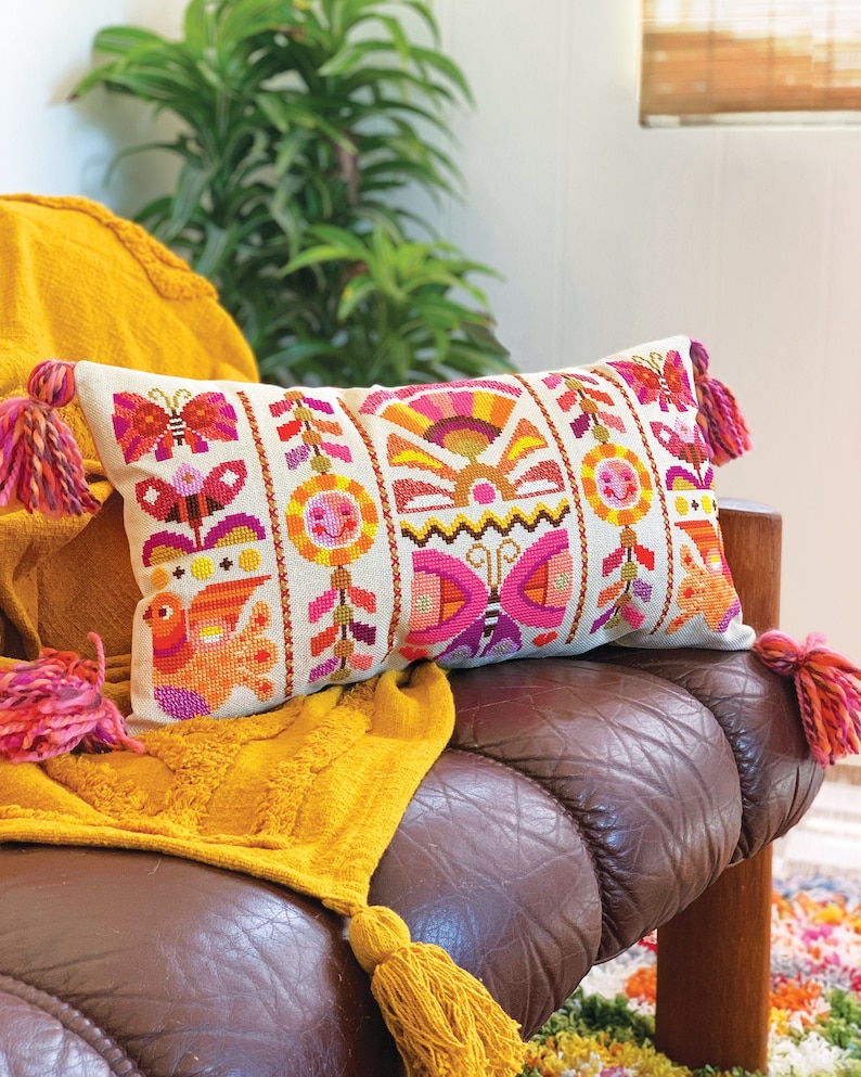 May include: A colourful embroidered cushion with a geometric pattern featuring butterflies, flowers, and suns. The cushion has tassels on each end and is resting on a brown leather sofa.