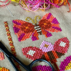 May include: Close-up of a cross-stitch embroidery project featuring two butterflies. The butterflies are stitched in shades of orange, pink, and brown. A pair of black embroidery scissors are in the foreground.
