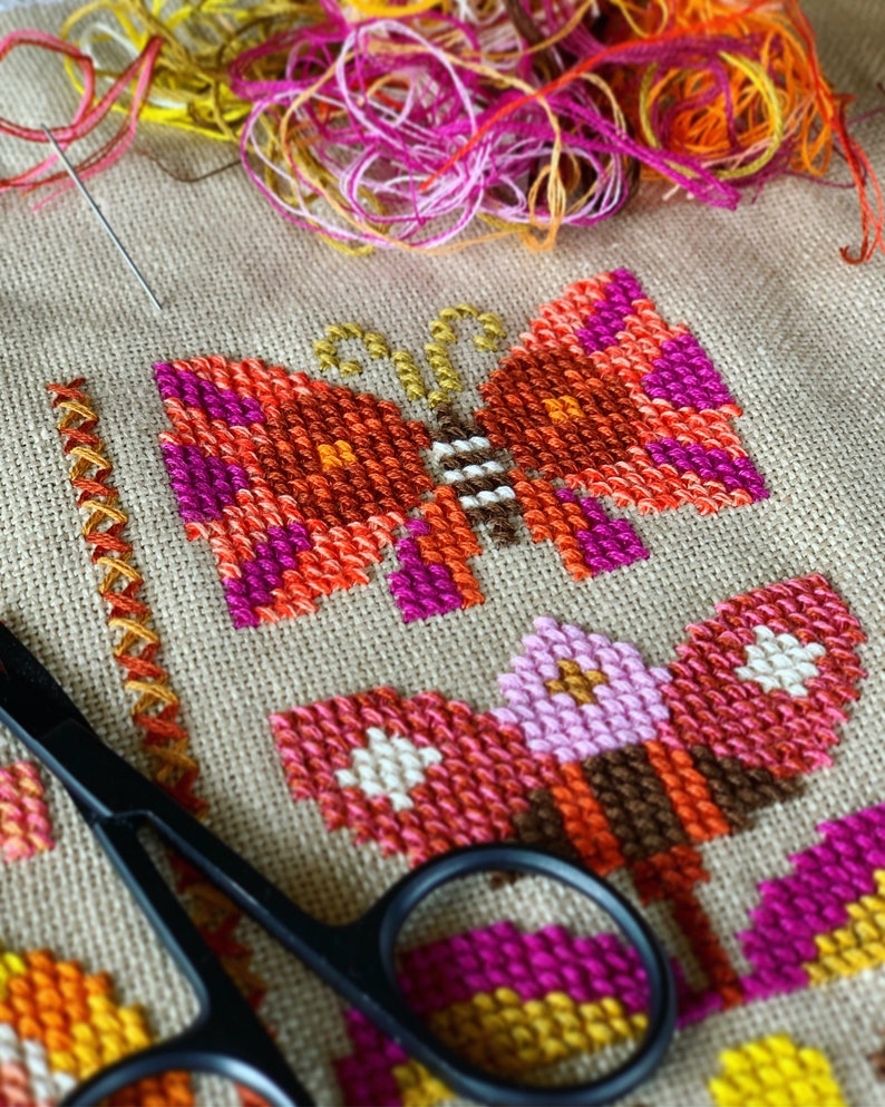 May include: Close-up of a cross-stitch embroidery project featuring two butterflies. The butterflies are stitched in shades of orange, pink, and brown. A pair of black embroidery scissors are in the foreground.