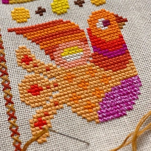 May include: Close-up of a cross-stitch embroidery project featuring a colourful bird with orange, yellow, pink, and purple thread. The needle and thread are visible in the bottom right corner of the image.