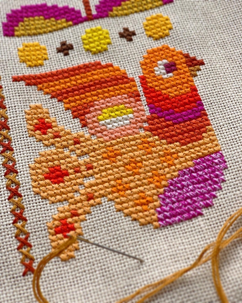 May include: Close-up of a cross-stitch embroidery project featuring a colourful bird with orange, yellow, pink, and purple thread. The needle and thread are visible in the bottom right corner of the image.
