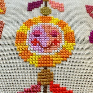 May include: A cross-stitch pattern of a smiling flower with orange, yellow, pink, and red accents. The flower is hanging from a stem with brown, orange, and red leaves.
