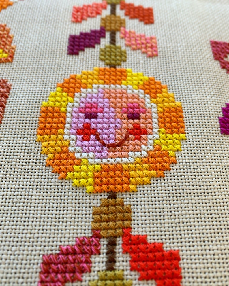 May include: A cross-stitch pattern of a smiling flower with orange, yellow, pink, and red accents. The flower is hanging from a stem with brown, orange, and red leaves.