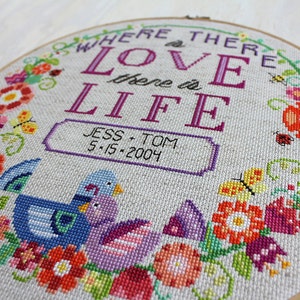 K&ouml;nnte beinhalten: Ein Kreuzstich-Stickrahmen mit einem floralen Kranzdesign. In der Mitte befindet sich der Text "Where there is love there is life" in lilafarbener Schrift, darunter ein kleineres Banner mit der Aufschrift "Jess + Tom 5-15-2004". Zwei lila und blaue V&ouml;gel sind unter dem Banner gestickt.