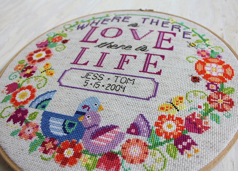 K&ouml;nnte beinhalten: Ein Kreuzstich-Stickrahmen mit einem floralen Kranzdesign. In der Mitte befindet sich der Text "Where there is love there is life" in lilafarbener Schrift, darunter ein kleineres Banner mit der Aufschrift "Jess + Tom 5-15-2004". Zwei lila und blaue V&ouml;gel sind unter dem Banner gestickt.