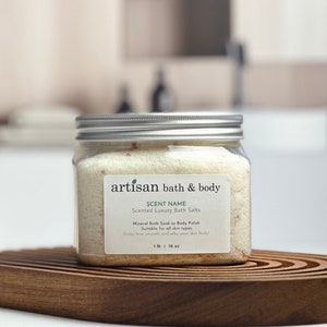 May include: A clear glass jar with a silver lid filled with white bath salts. The jar has a label that reads "artisan bath & body" and "SCENT NAME Scented Luxury Bath Salts". The label also says "Mineral Bath Soak or Body Polish Suitable for all skin types".