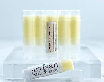 May include: Five clear plastic tubes of pale yellow arnica infused lip balm stand upright, with one additional tube lying horizontally in front. The labels feature the text artisan bath & body and Arnica Infused Lip Balm Natural Gloss.