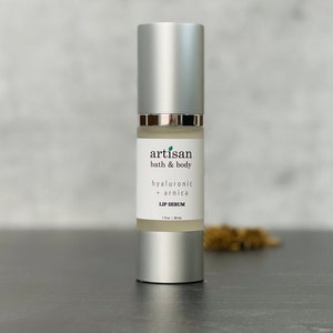 May include: A silver bottle with a white label that reads "artisan bath & body hyaluronic + arnica LIP SERUM 1 fl oz / 30 ml".